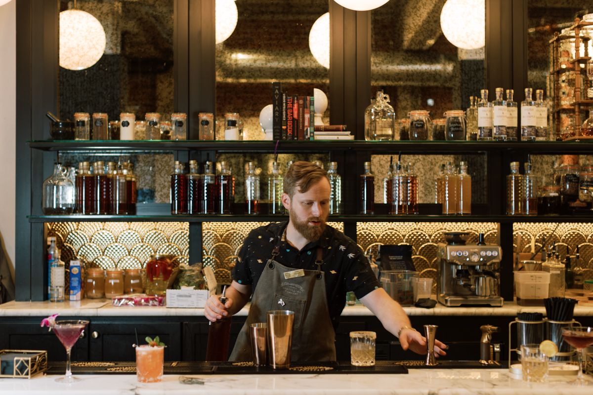 Bartender in apron mixing cocktails behind a stylish bar with bottle-lined shelves and golden mosaic tiles.