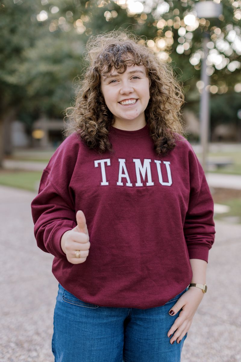 A curly-haired student gives a thumbs up wearing a maroon TAMU crewneck sweatshirt.