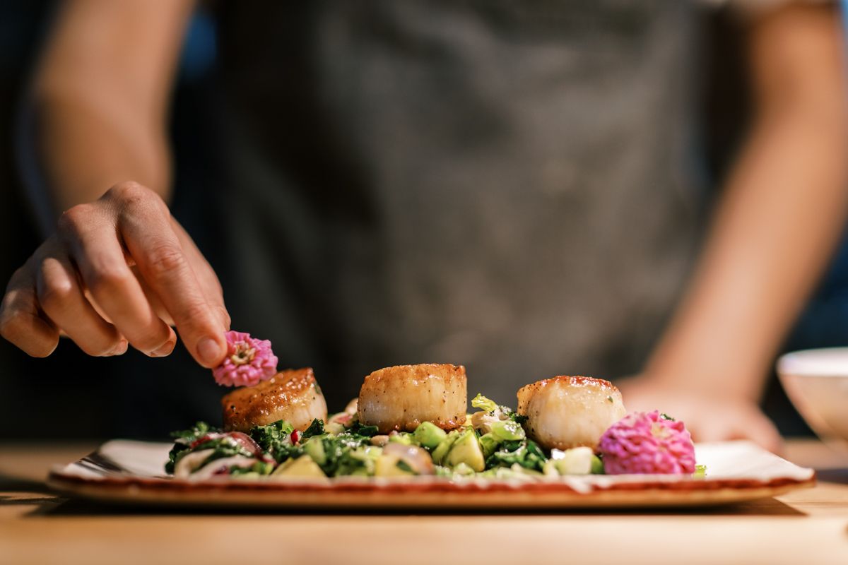 Chef plating seared scallops with greens and beet garnish on a white rectangular dish.