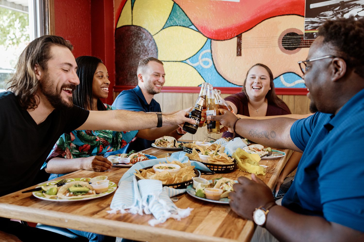 Diverse group of friends toasting drinks over a table full of food at a colorful Mexican restaurant.
