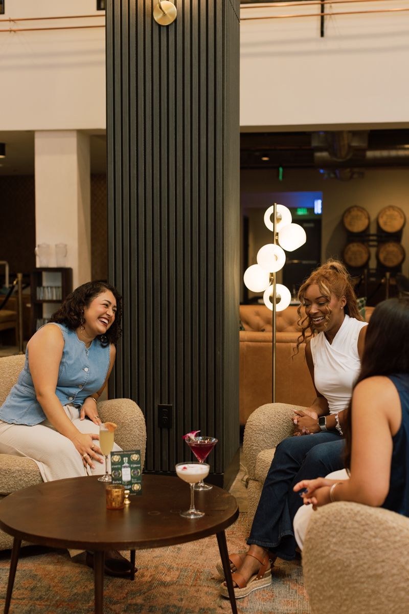 Three women laughing and socializing over cocktails in a modern distillery lounge with warm ambient lighting.