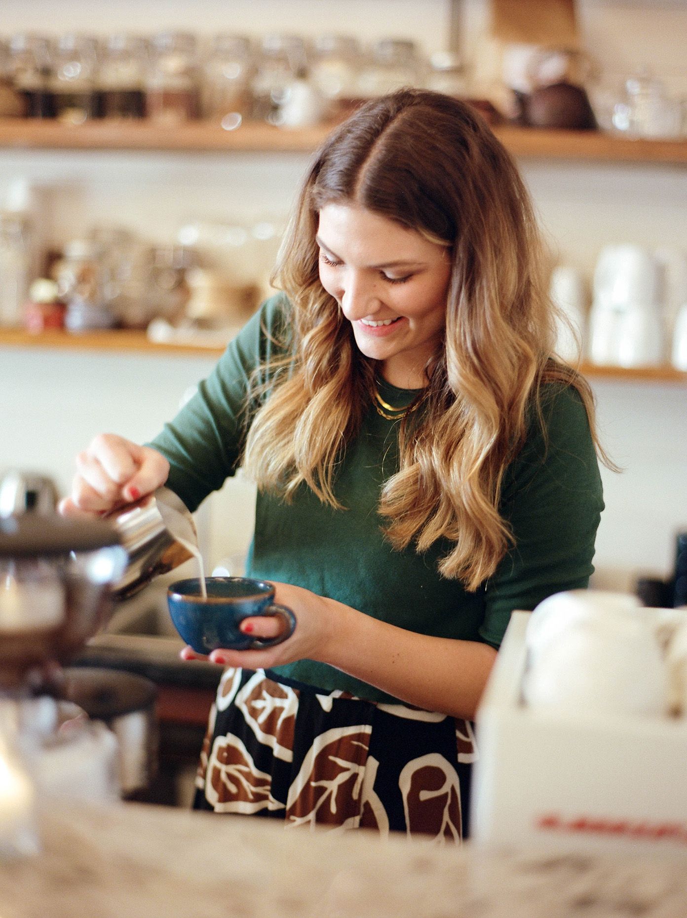 Smiling woman with long blonde hair pouring milk into a blue coffee cup in a cozy cafe kitchen.