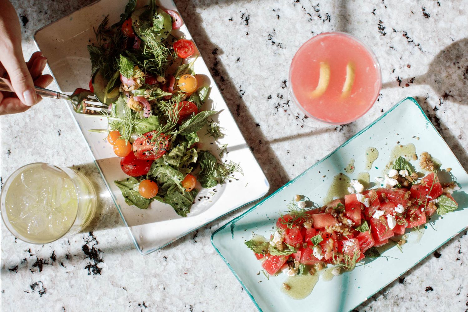 Overhead view of fresh tomato salads, pink cocktail, and lemon drink on a speckled white surface.