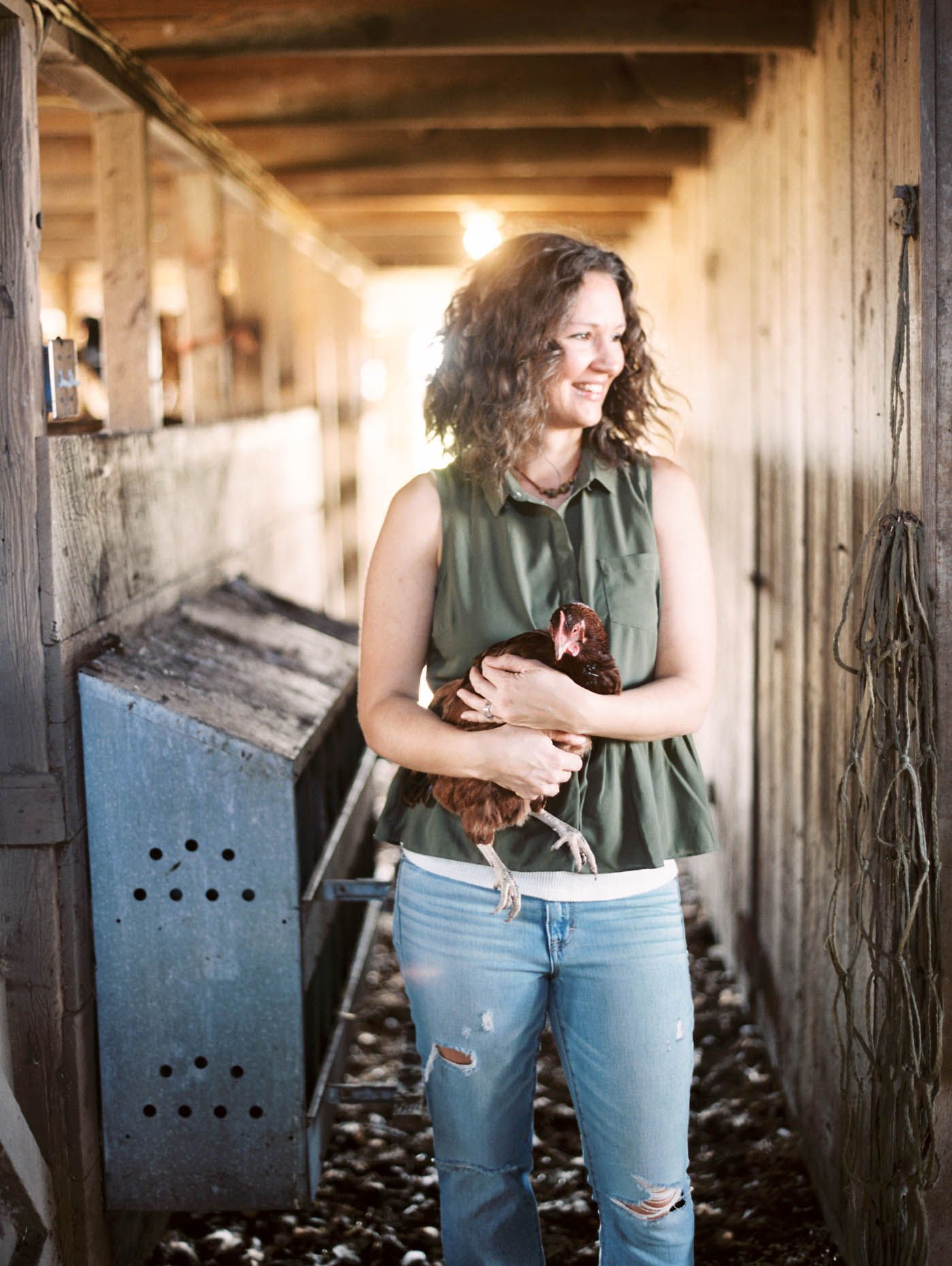 Woman in green top and jeans holds a brown chicken inside a rustic wooden barn corridor.