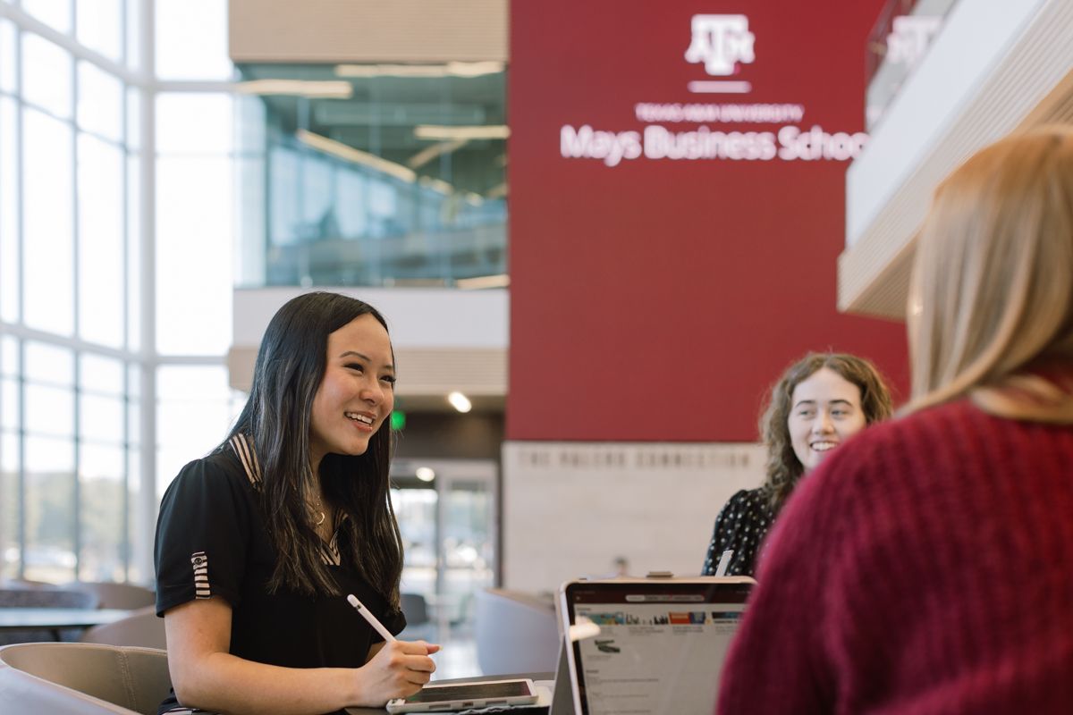A smiling student at Texas A&M Mays Business School works on a laptop at a reception desk.