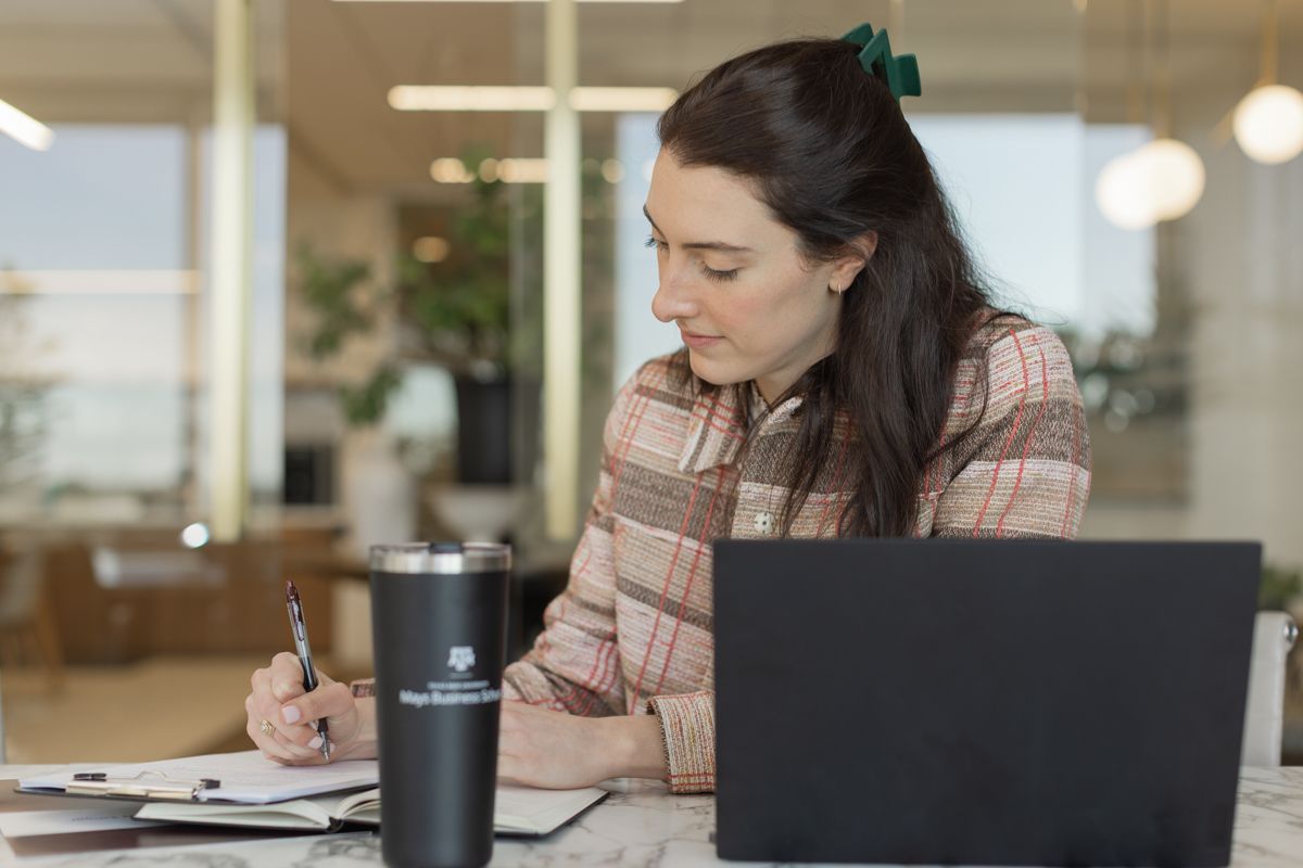Young woman taking notes in notebook beside laptop with branded tumbler in modern office space.