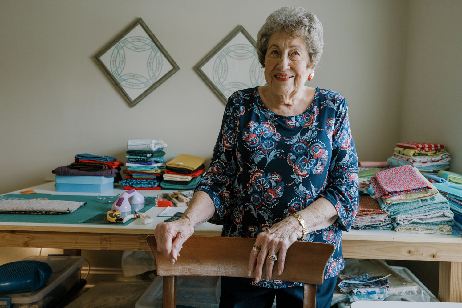 Elderly woman smiling at quilting table with colorful fabric stacks and framed quilts on wall behind her.