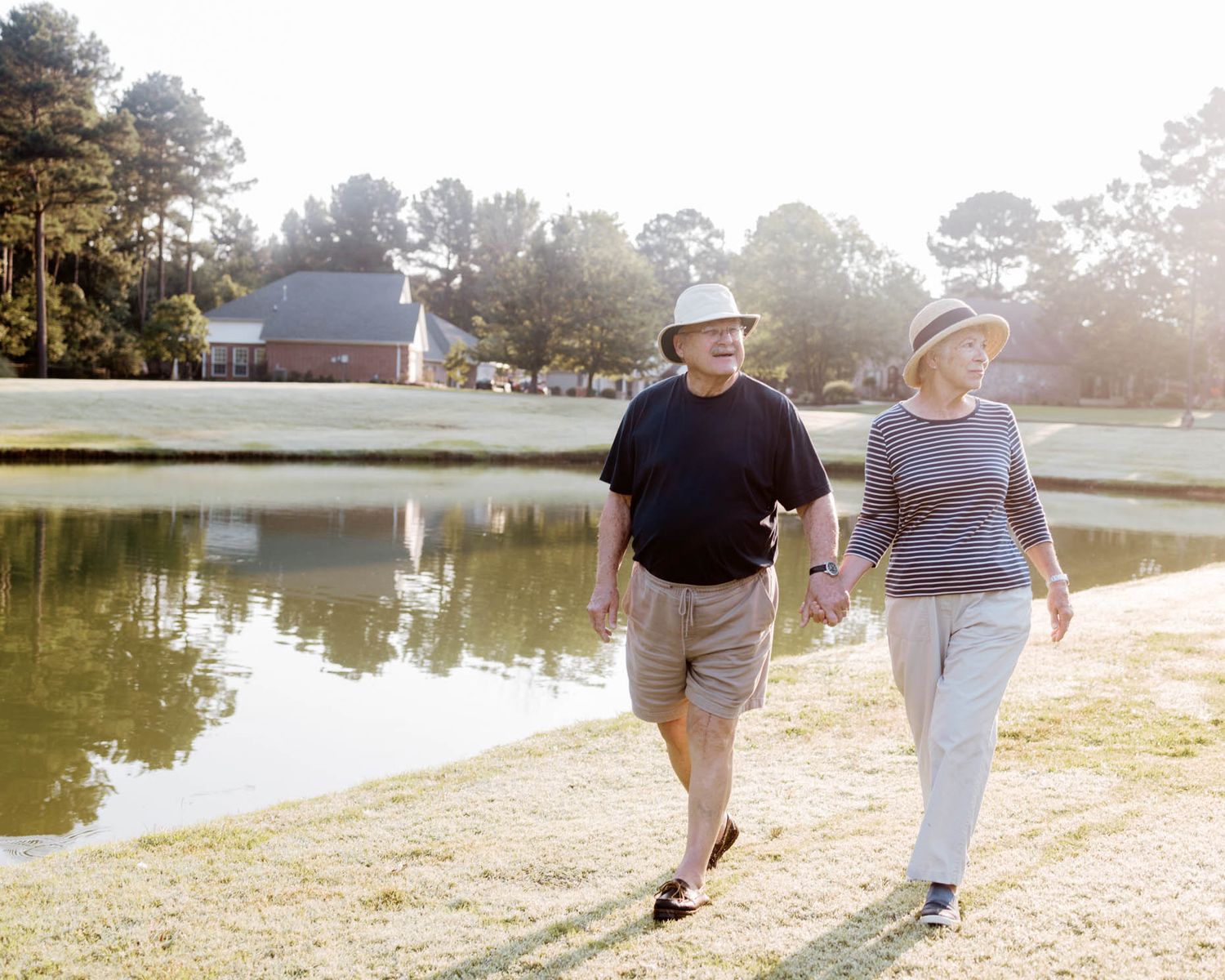 Elderly couple holding hands walking by a serene pond surrounded by trees on a sunny day.