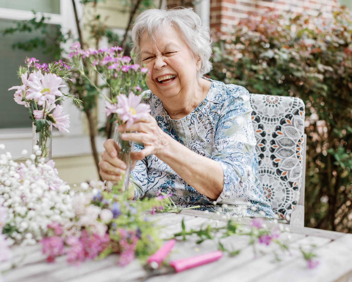 Elderly woman laughing joyfully while arranging fresh flowers at an outdoor table.