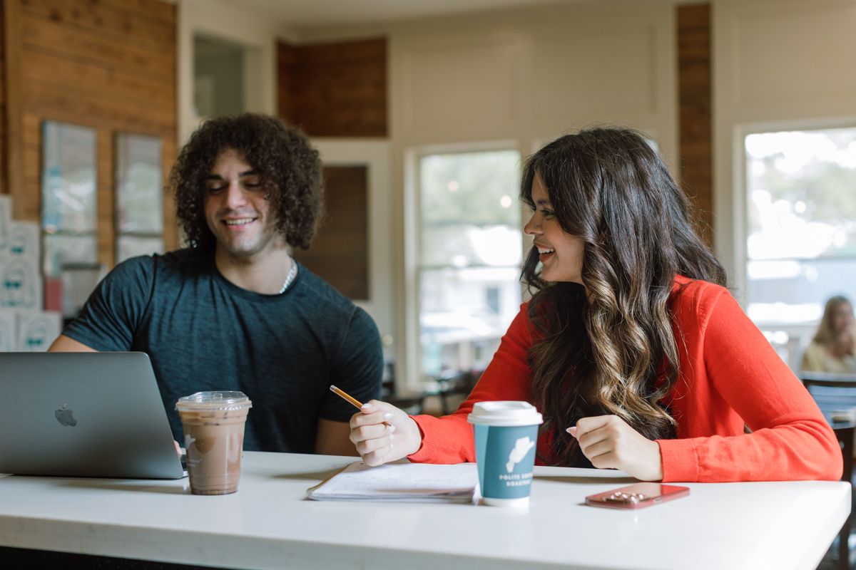 Two college students smiling and studying together on laptop at cafe with iced coffee drinks.