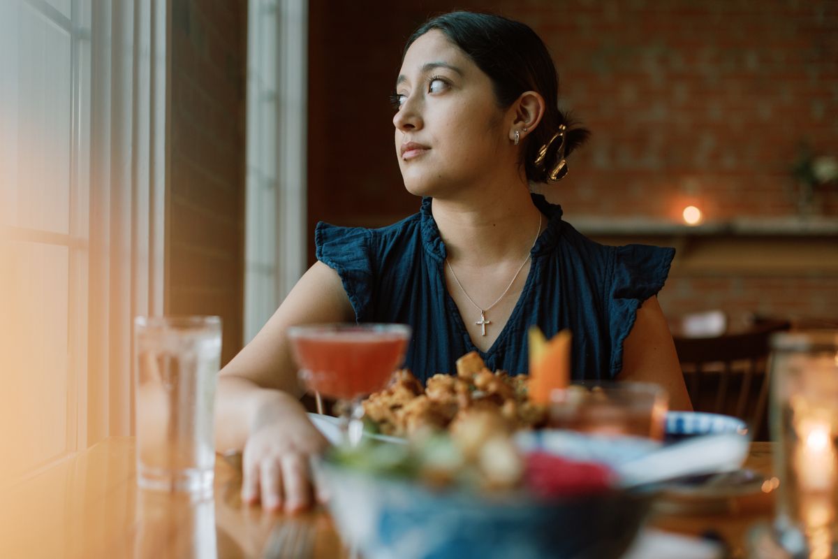 Young woman gazing out window at restaurant table with cocktail and appetizers in foreground.