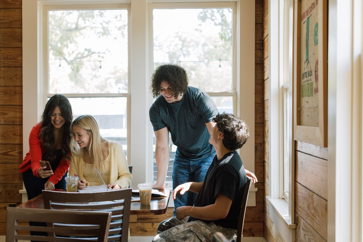 Four young friends laughing and socializing at a wooden table inside a cozy cafe restaurant.