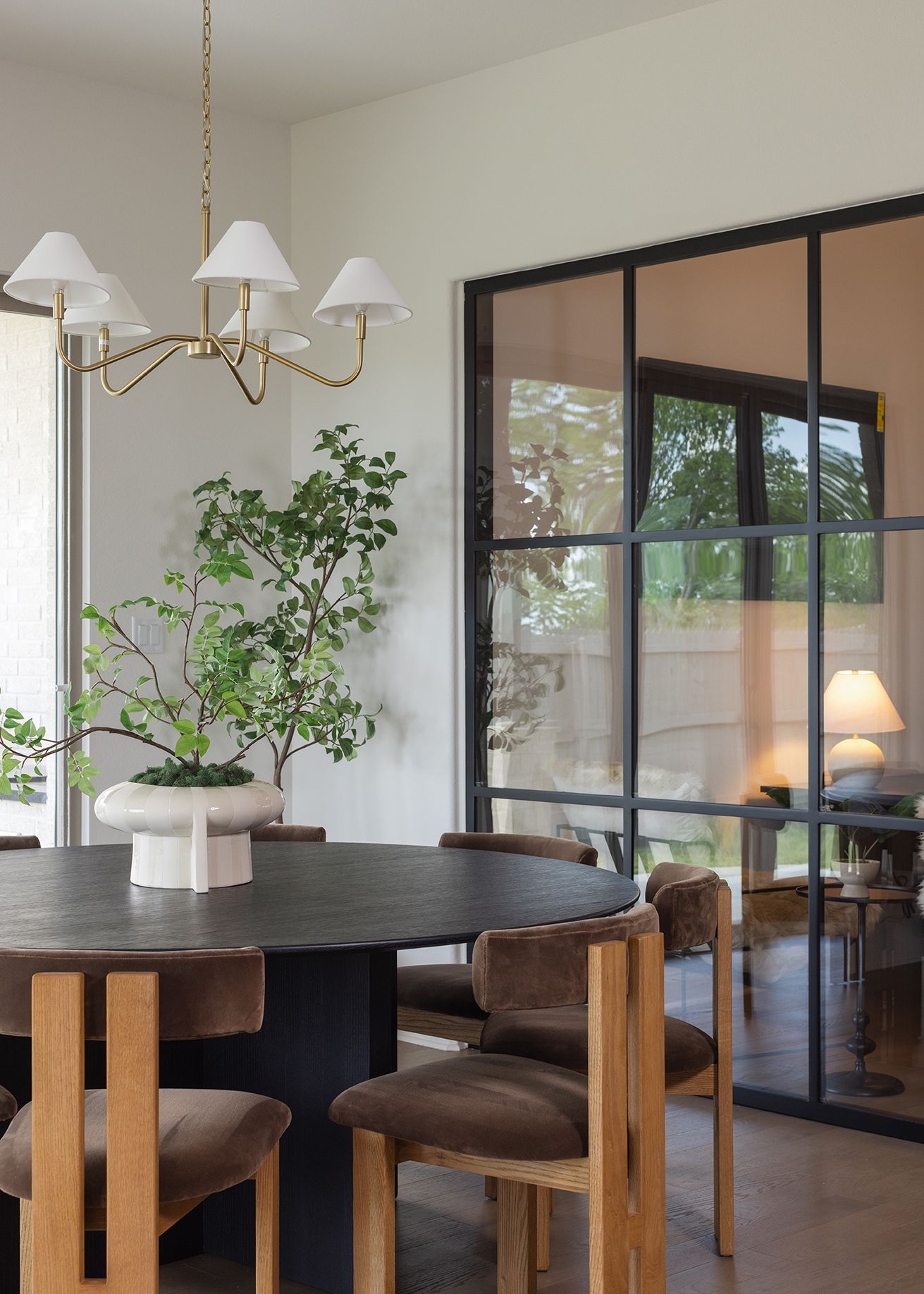 Modern dining area with black oval table, velvet chairs, brass chandelier, and steel-framed glass partition doors.
