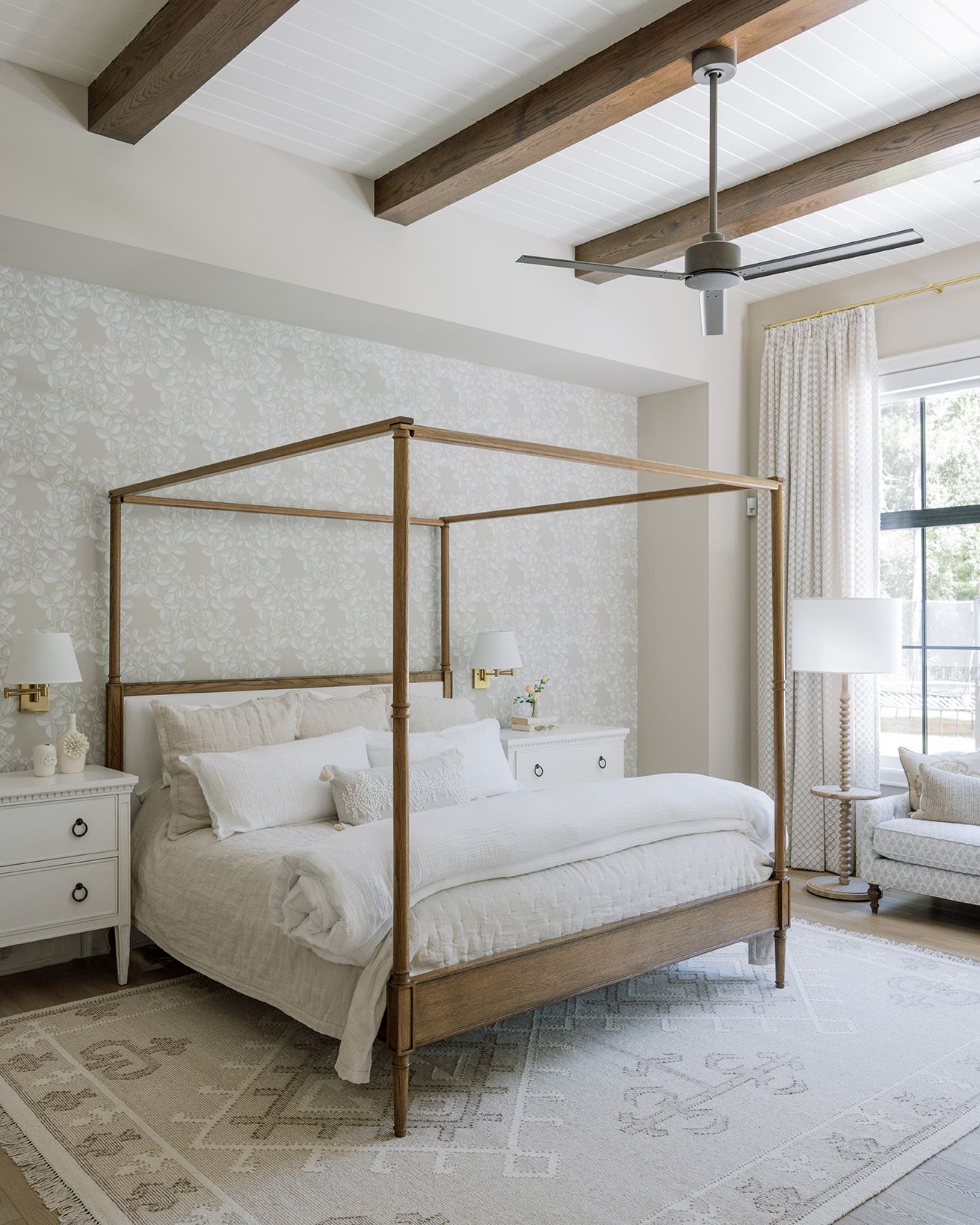 Elegant primary bedroom with four-poster bed, exposed wood beams, floral wallpaper, and neutral layered textiles.