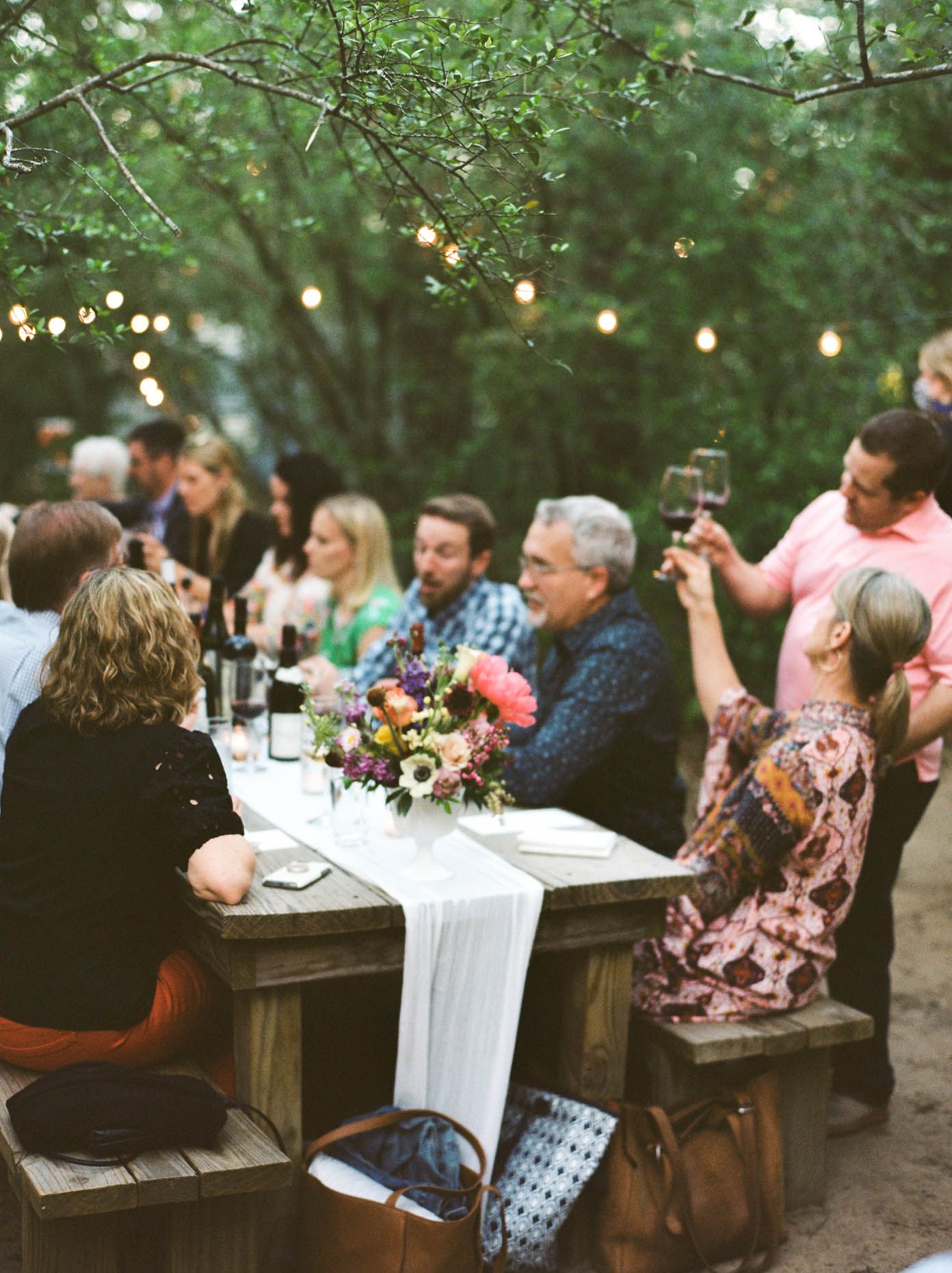Guests enjoy an outdoor dinner party at rustic wooden tables adorned with colorful flowers under string lights in a wooded setting.