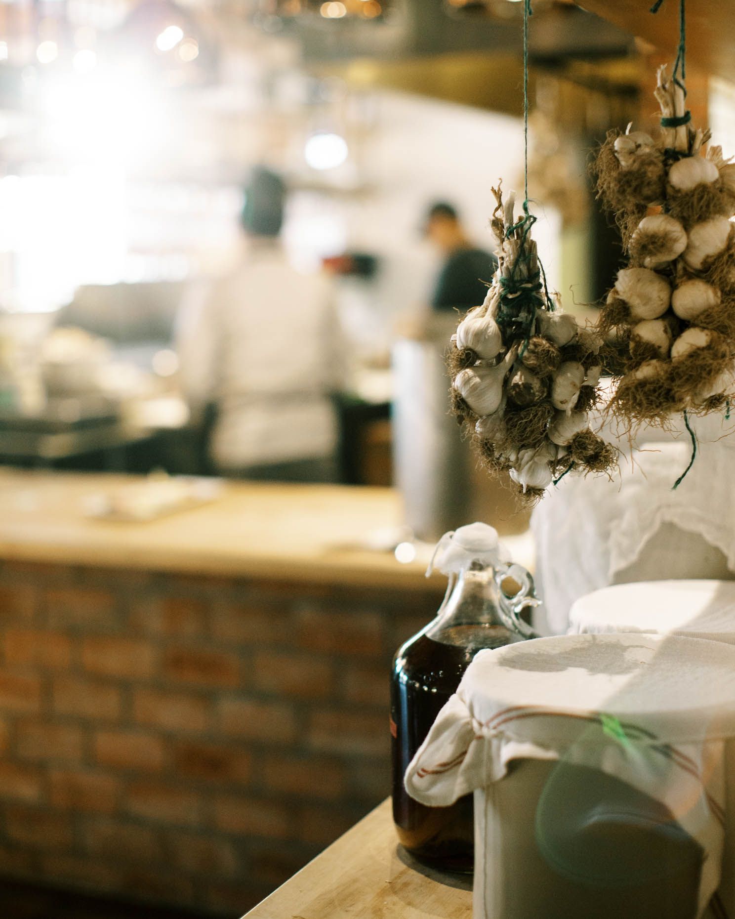 Rustic restaurant kitchen with hanging garlic bulbs, dark bottle, and chefs working in the warm background.