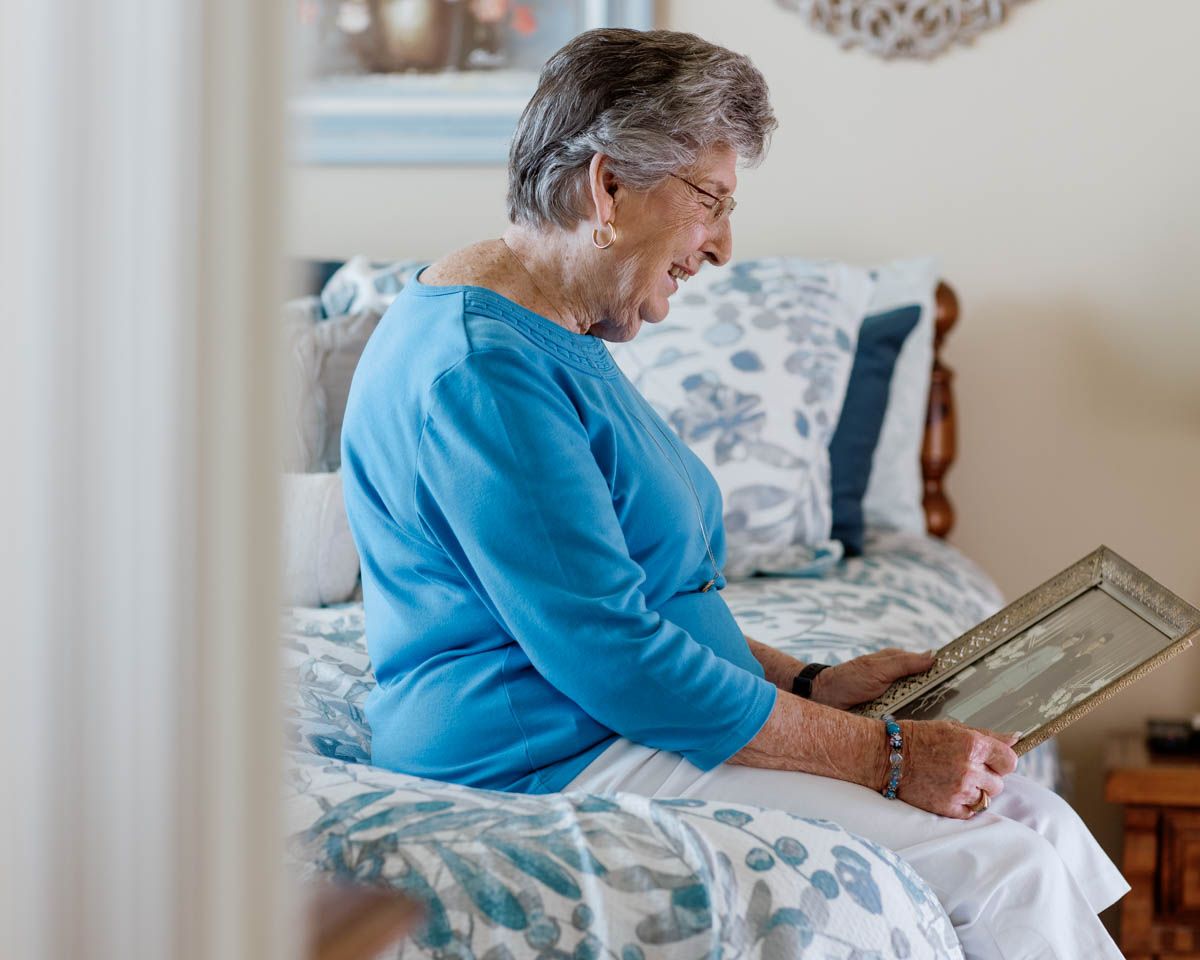 Elderly woman sitting on bed smiling while looking at a framed photo, wearing a blue top.