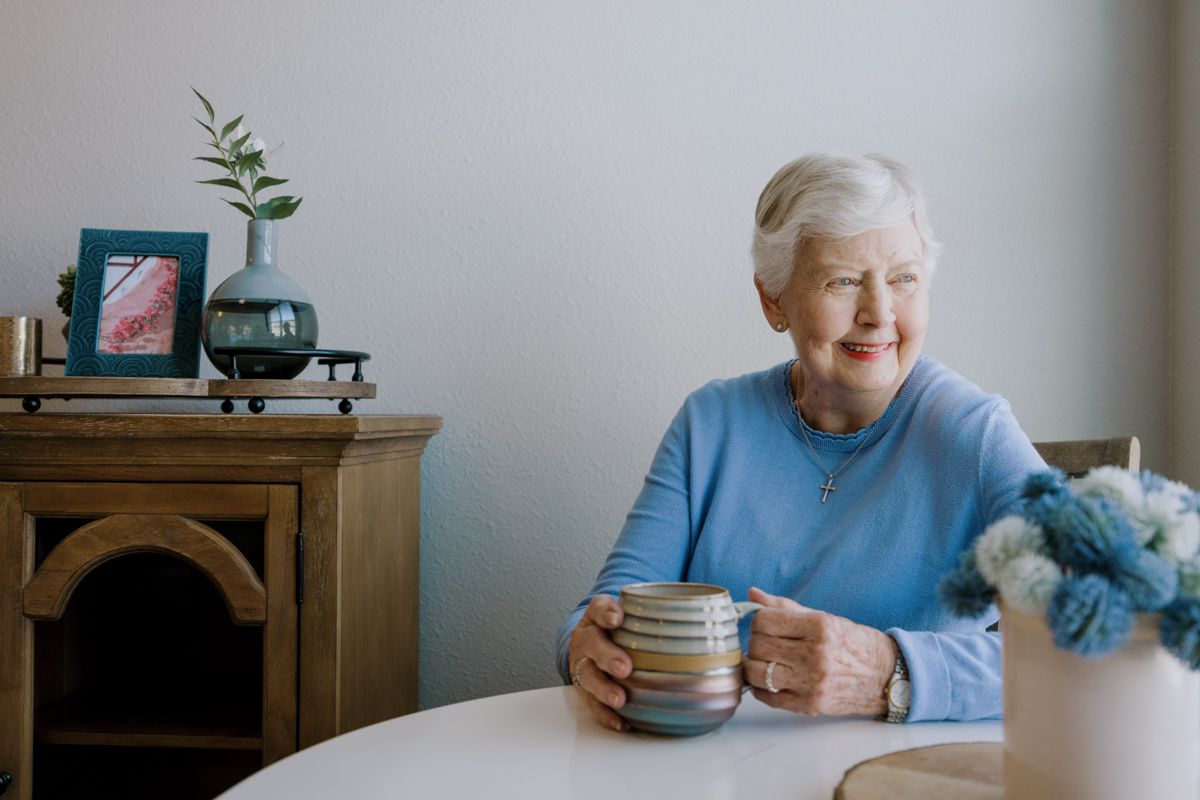 Elderly woman smiling while holding a striped mug at a white table in a cozy home setting.