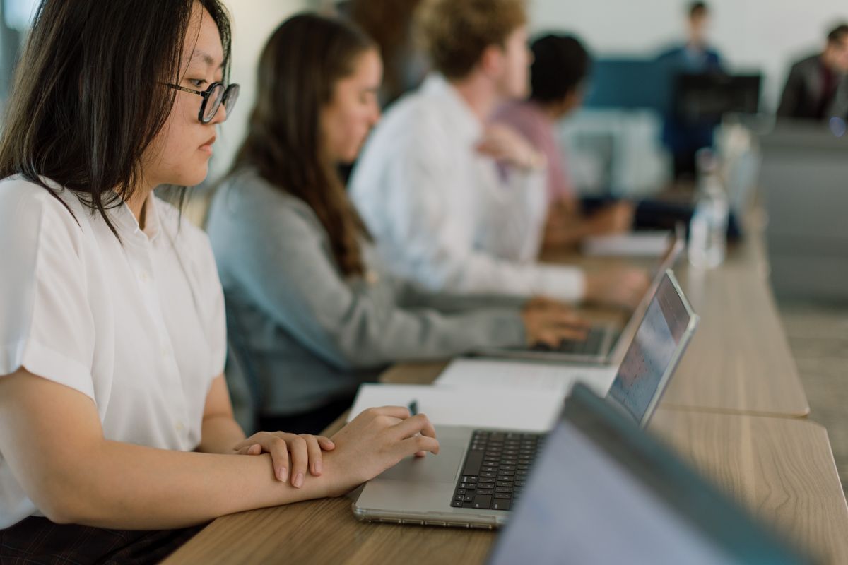 Students work on laptops at a long desk in a modern university classroom setting.