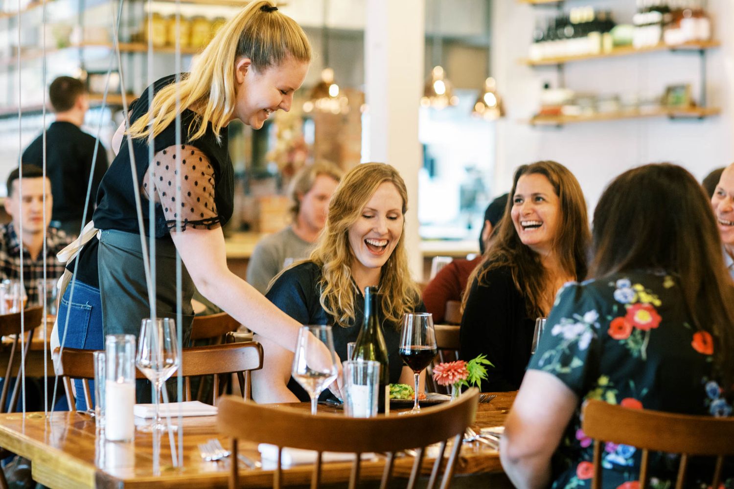 Waitress serving wine to laughing women at a busy restaurant dining table.