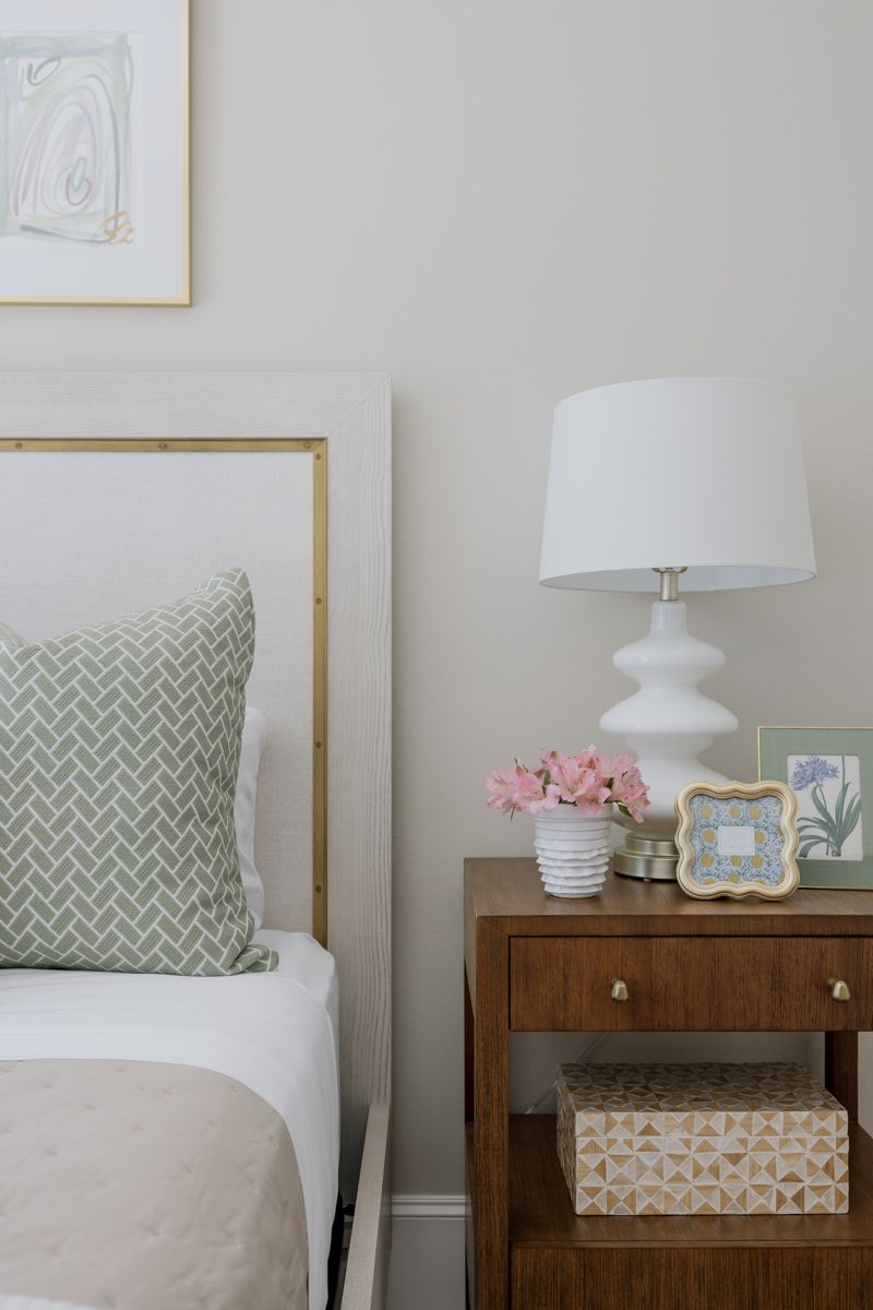 Cozy bedroom nightstand with white lamp, pink flowers, and green framed art beside a gold headboard bed.