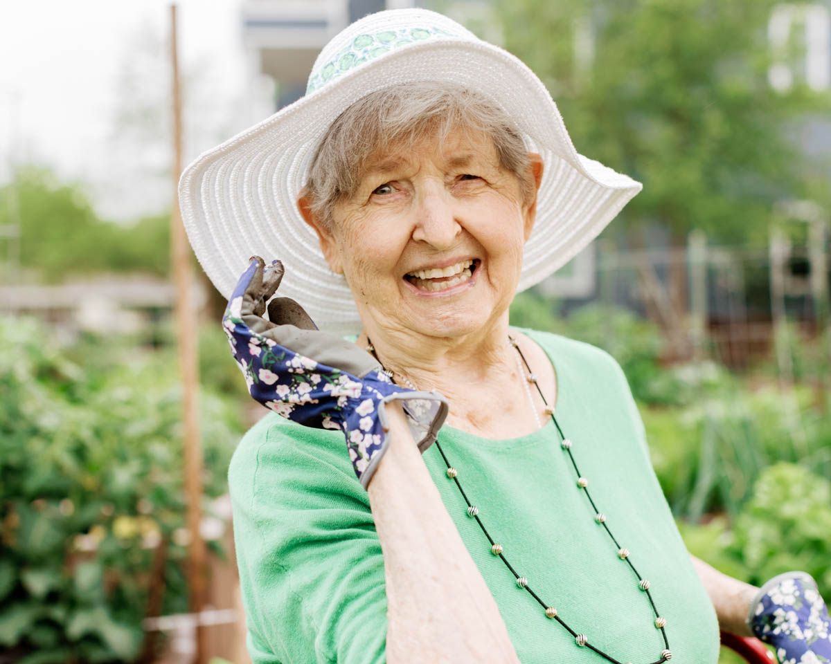 Cheerful senior woman in a white sun hat and gardening gloves smiling while working in a lush garden.