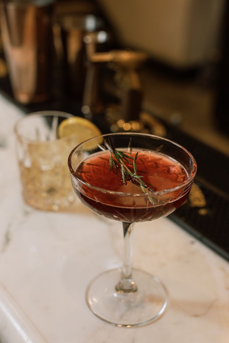 A dark red cocktail garnished with rosemary in a coupe glass on a marble bar top.