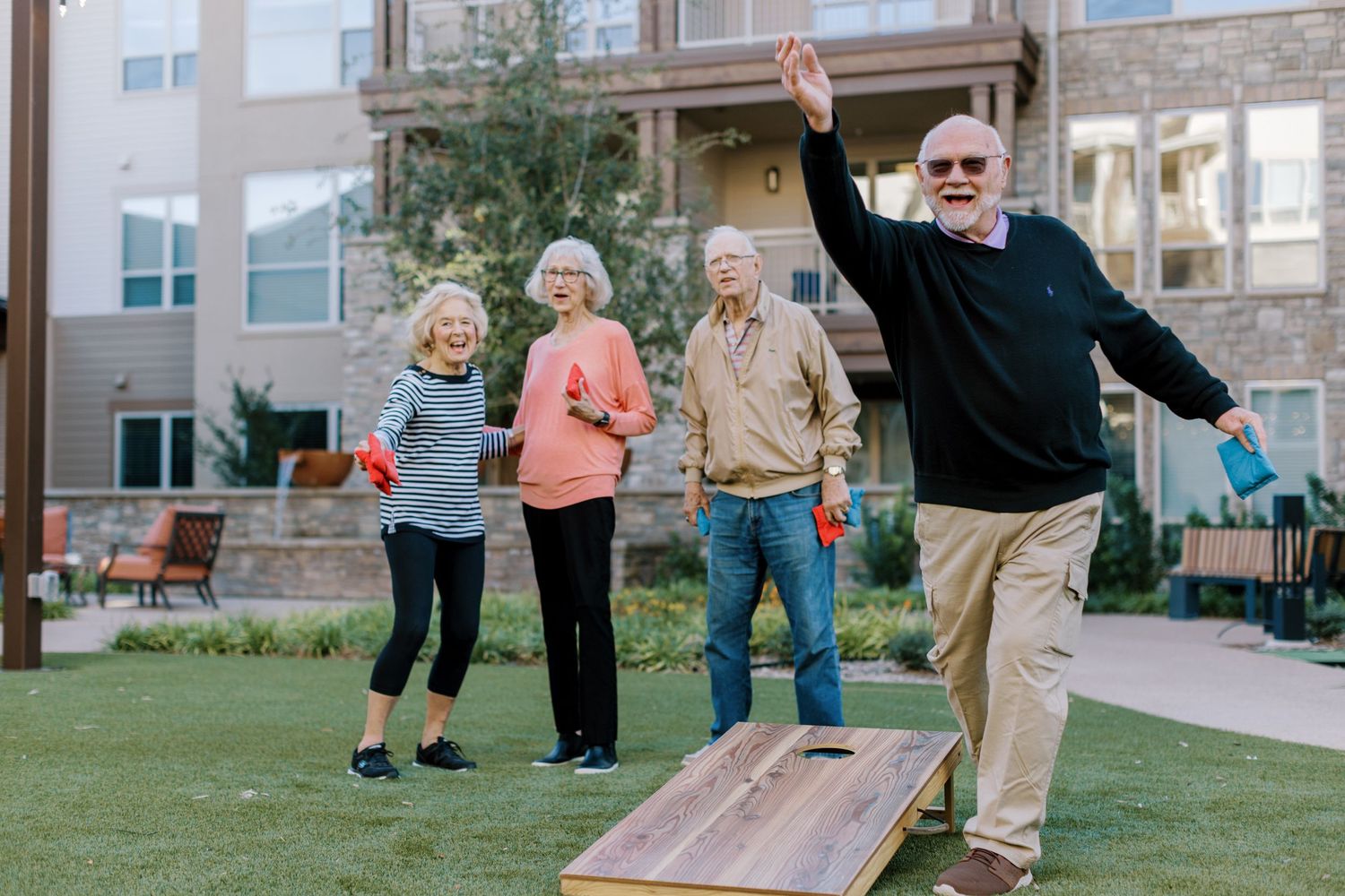 Four seniors playing cornhole on a green lawn outside a residential community building on a sunny day.