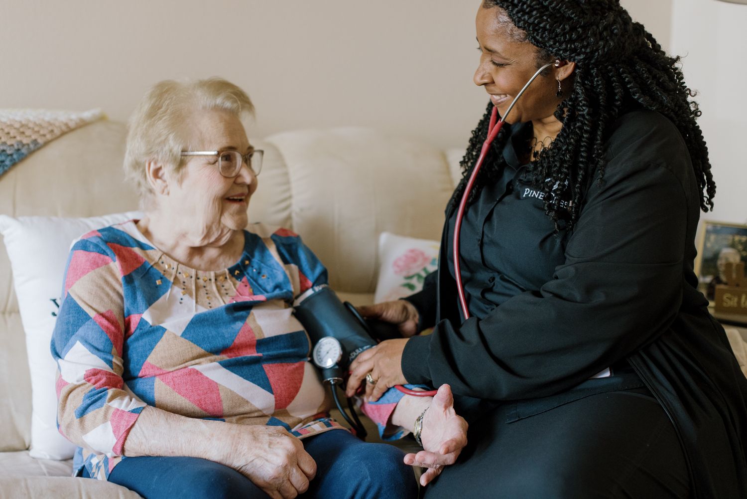 A Black female nurse uses a stethoscope to check an elderly white woman's health at home.