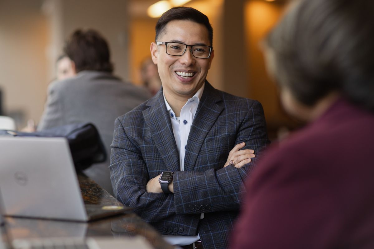 Smiling Asian businessman in plaid blazer sits with arms crossed near a laptop.
