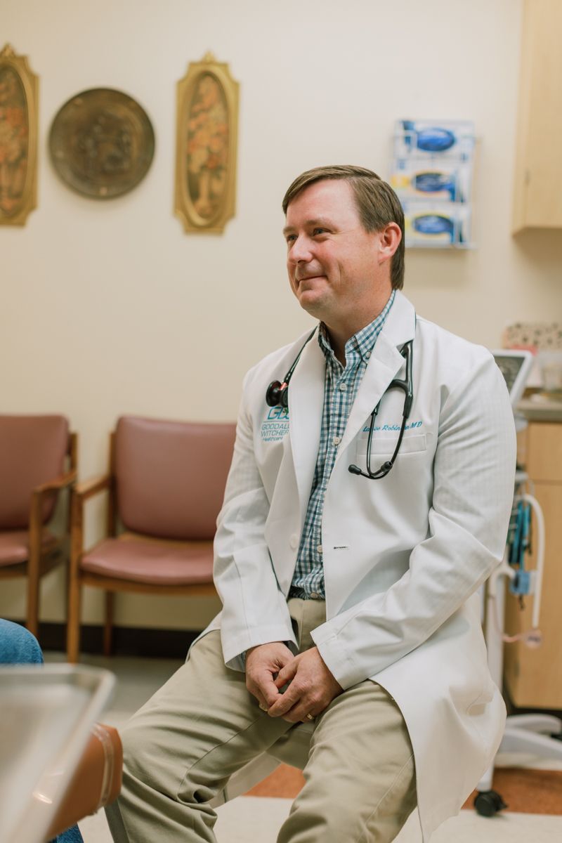 A smiling male doctor in a white coat with stethoscope sits in a medical examination room.