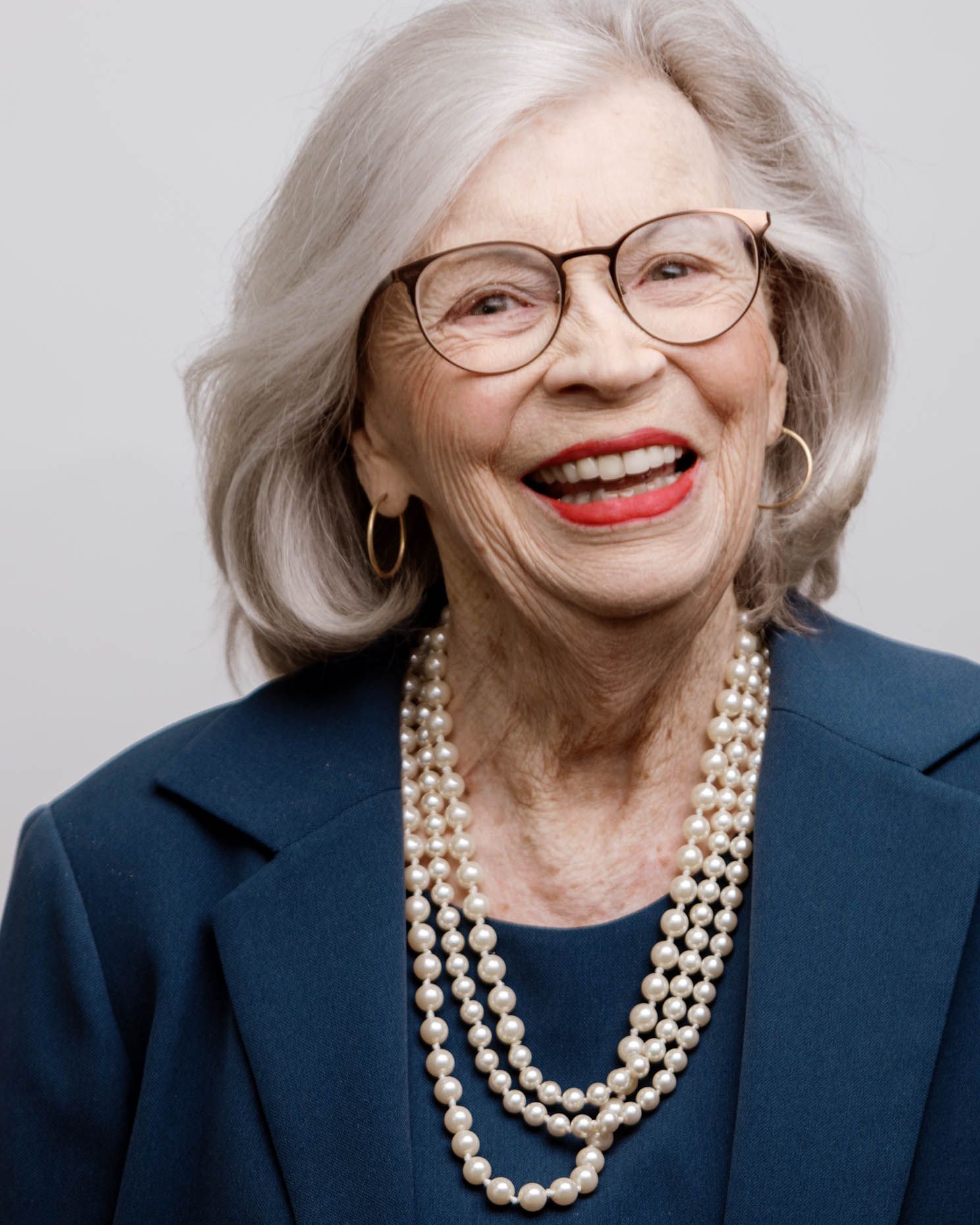 Elegant senior woman smiling confidently wearing pearl necklace, glasses, and a navy blazer.