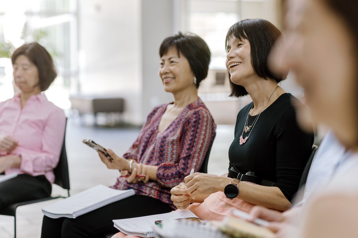 Smiling Asian women sitting in rows at a seminar, holding notebooks and phones, engaged in a presentation.