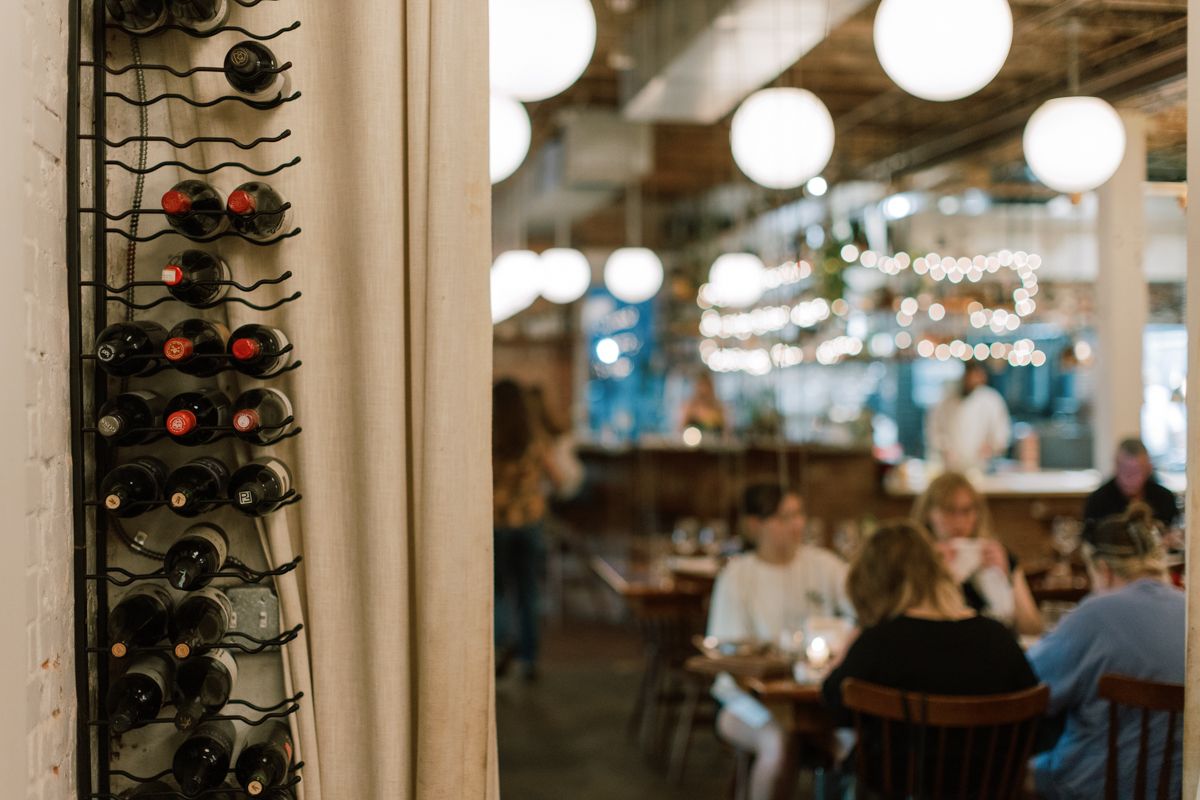 Wine rack with bottles near a warmly lit restaurant dining area with guests seated at tables.