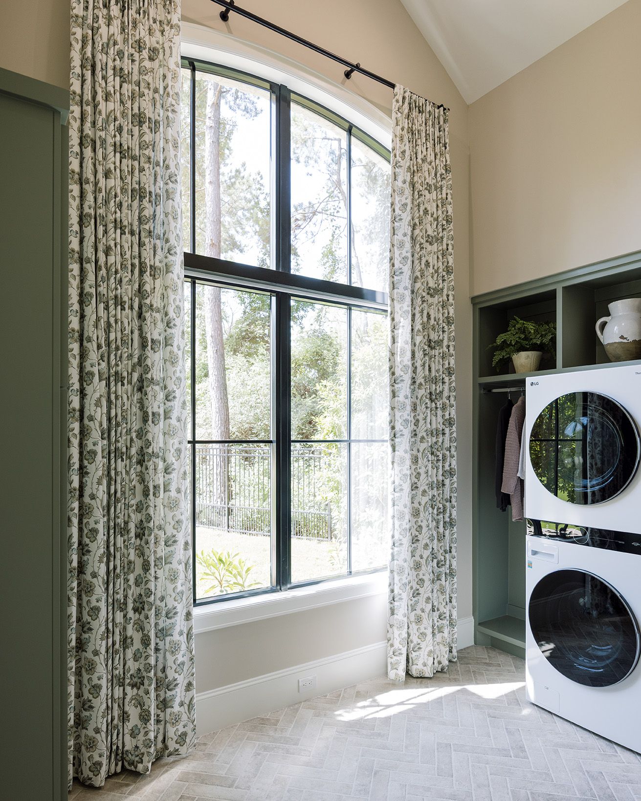 Bright laundry room with arched black-frame window, floral curtains, green built-ins, and stacked washer dryer unit.