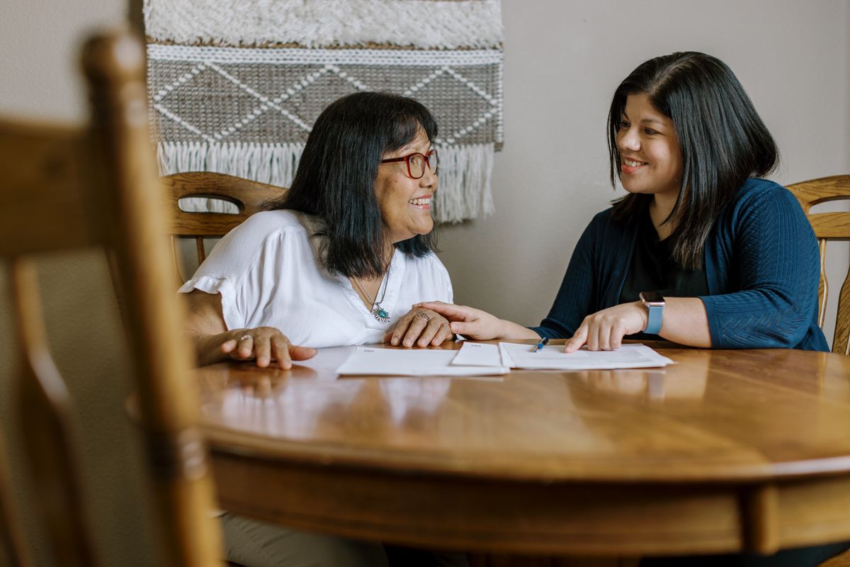 Two young women smiling at each other while sitting at a wooden dining table together.