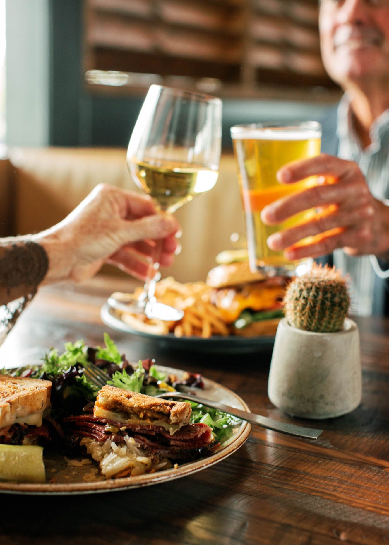 Two people toasting with white wine and beer over plates of sandwiches and fries at a restaurant table.