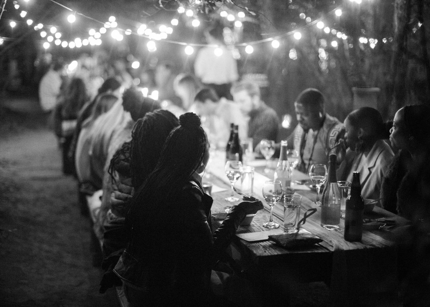 Guests dining at a long outdoor table under string lights at a nighttime garden dinner party.