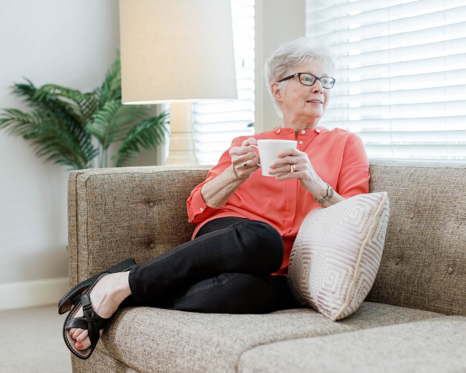 Elderly woman with glasses relaxing on a sofa, holding a white mug, with a plant and lamp in background.
