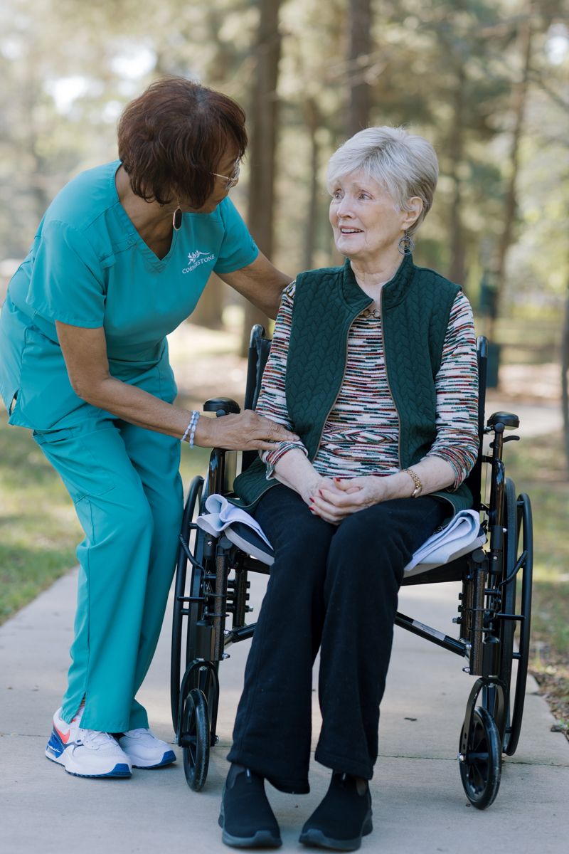 A nurse in teal scrubs assists an elderly woman in a wheelchair on an outdoor path surrounded by trees.