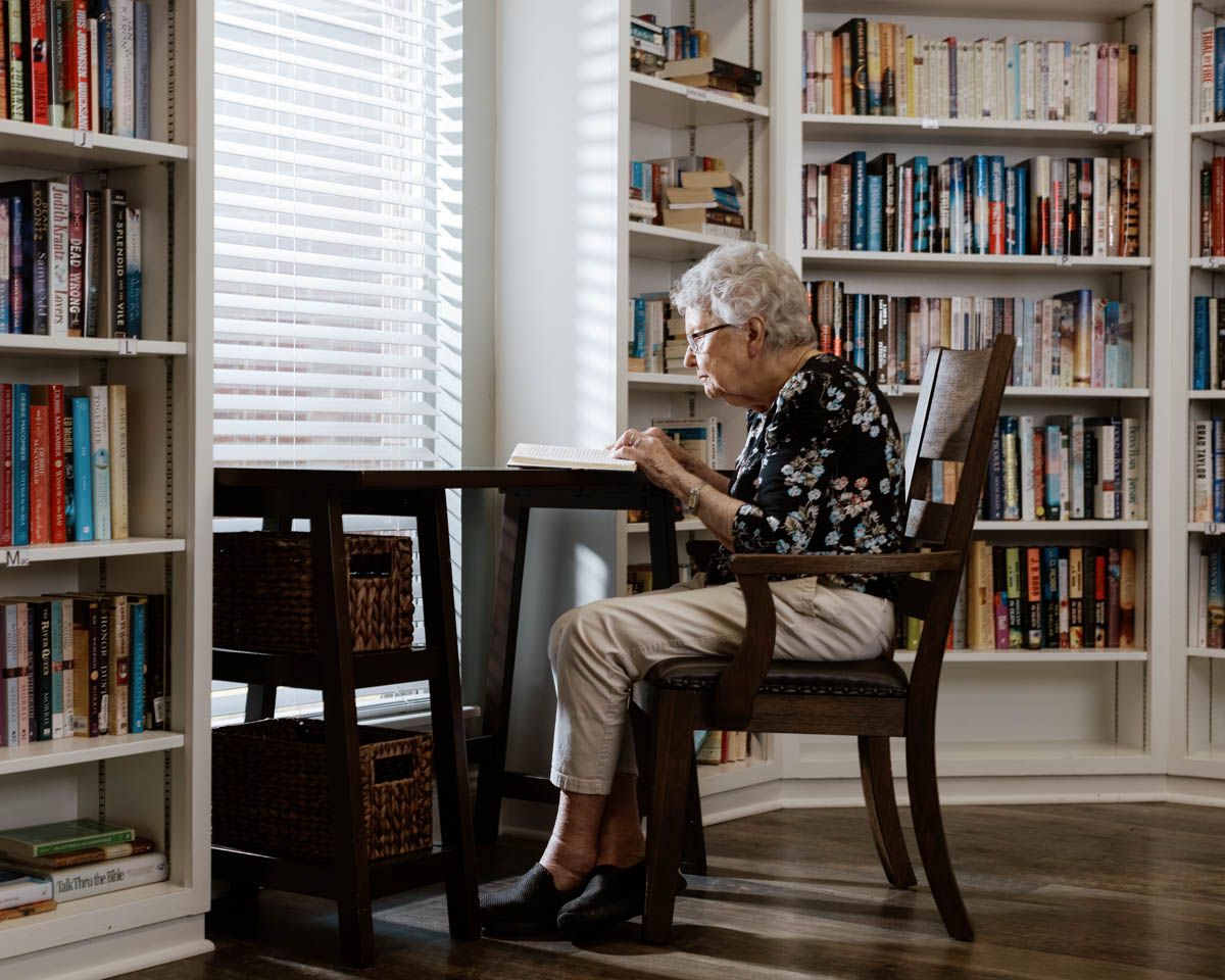 Elderly woman sitting alone at a wooden table in a cozy home library surrounded by bookshelves near a bright window.
