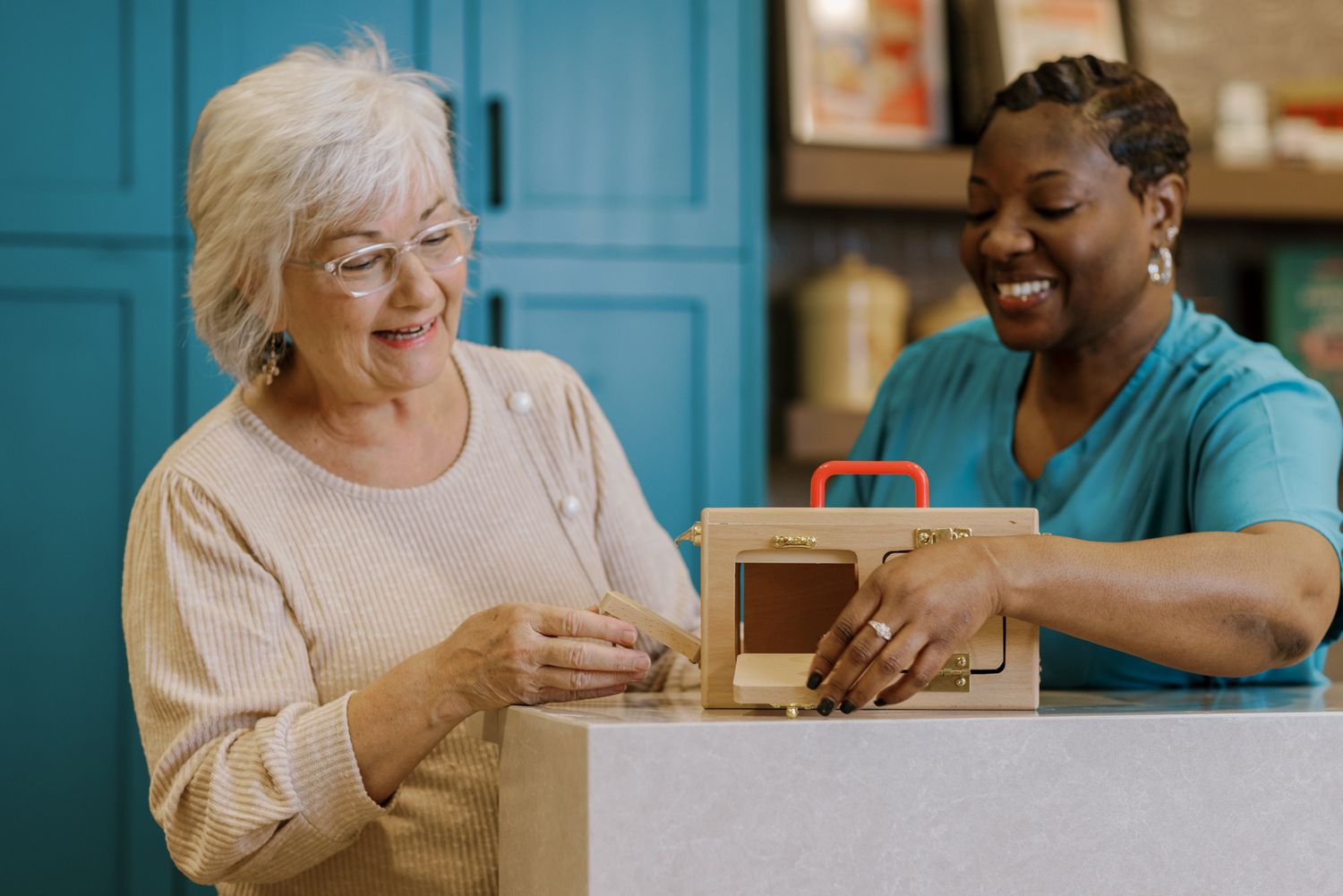 Two women smiling while opening a cardboard box together indoors.