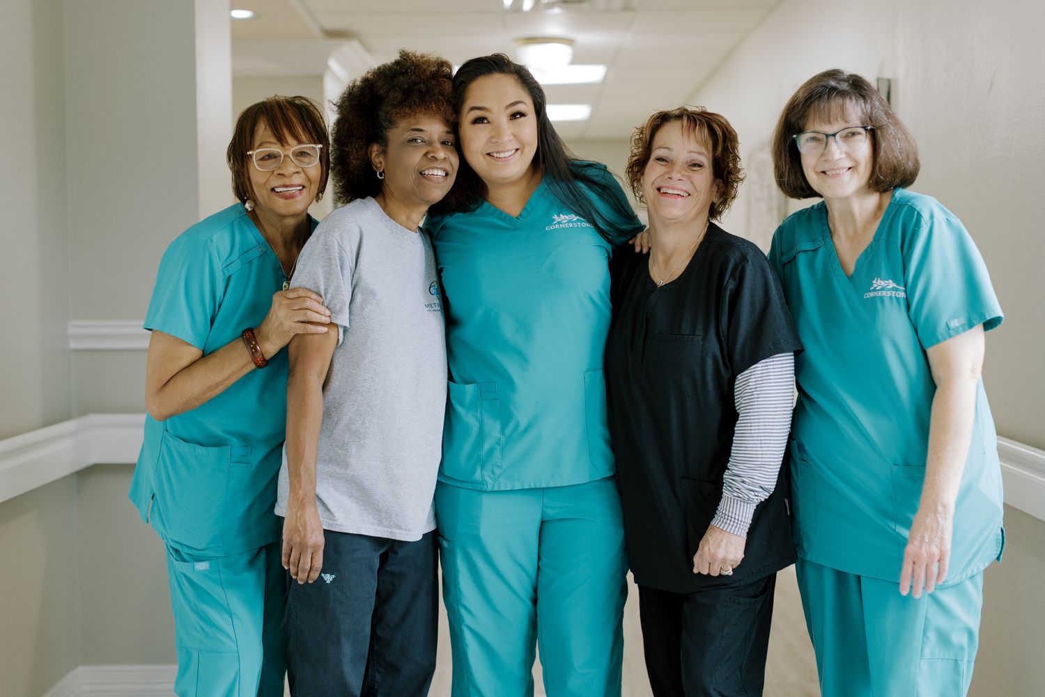 Five smiling healthcare workers in teal scrubs pose together in a bright hospital hallway.