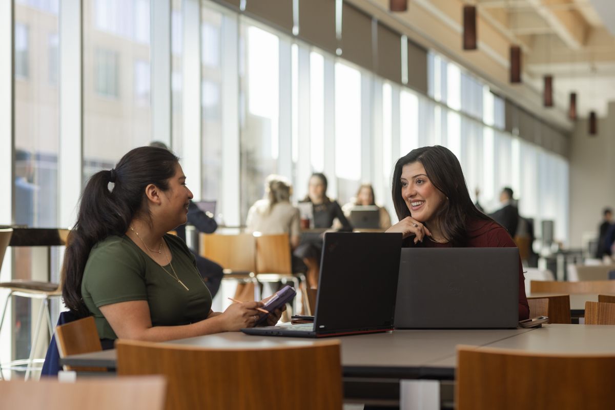 Two young women having a conversation at a table with a laptop in a bright modern space.