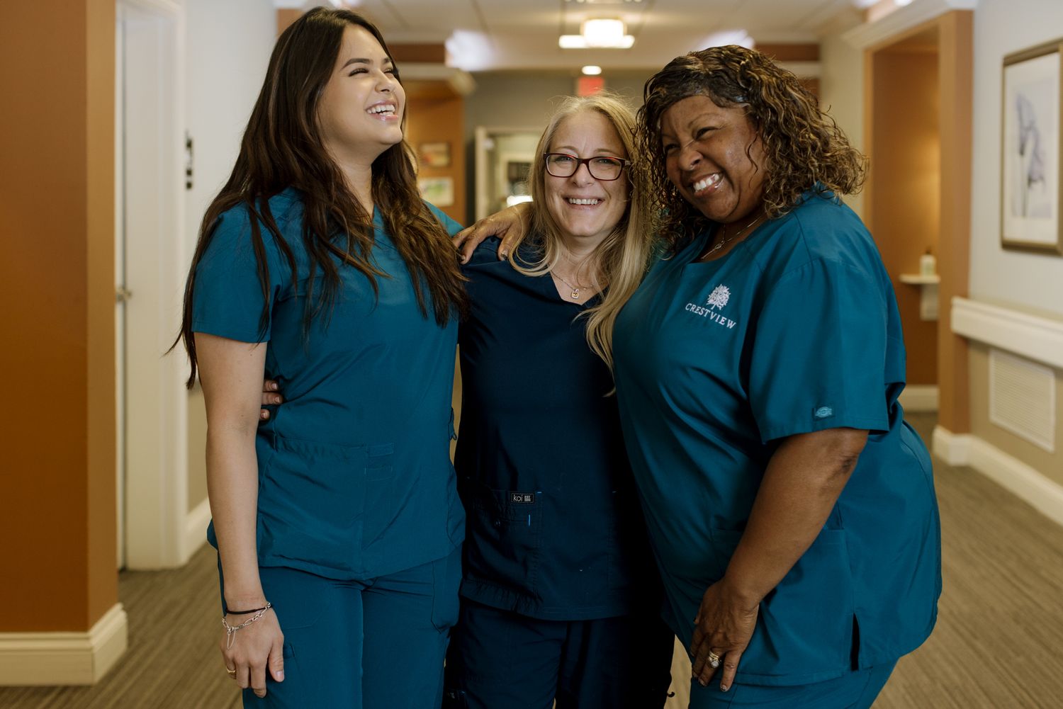 Three female healthcare workers in teal scrubs smiling together in a bright hospital hallway.