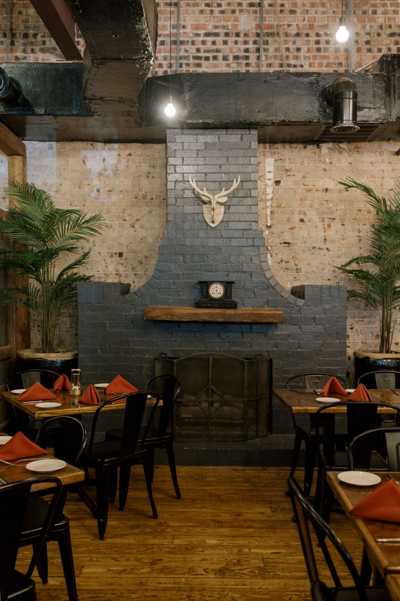 Rustic restaurant interior with dark brick fireplace, deer antler wall mount, black metal chairs, and red napkins on wooden tables.
