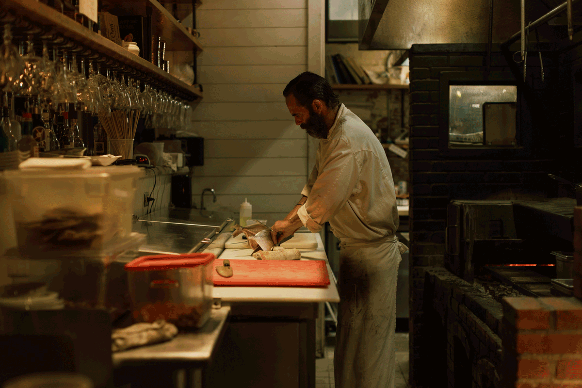 Chef preparing food in a professional restaurant kitchen with brick oven and wine glasses overhead.