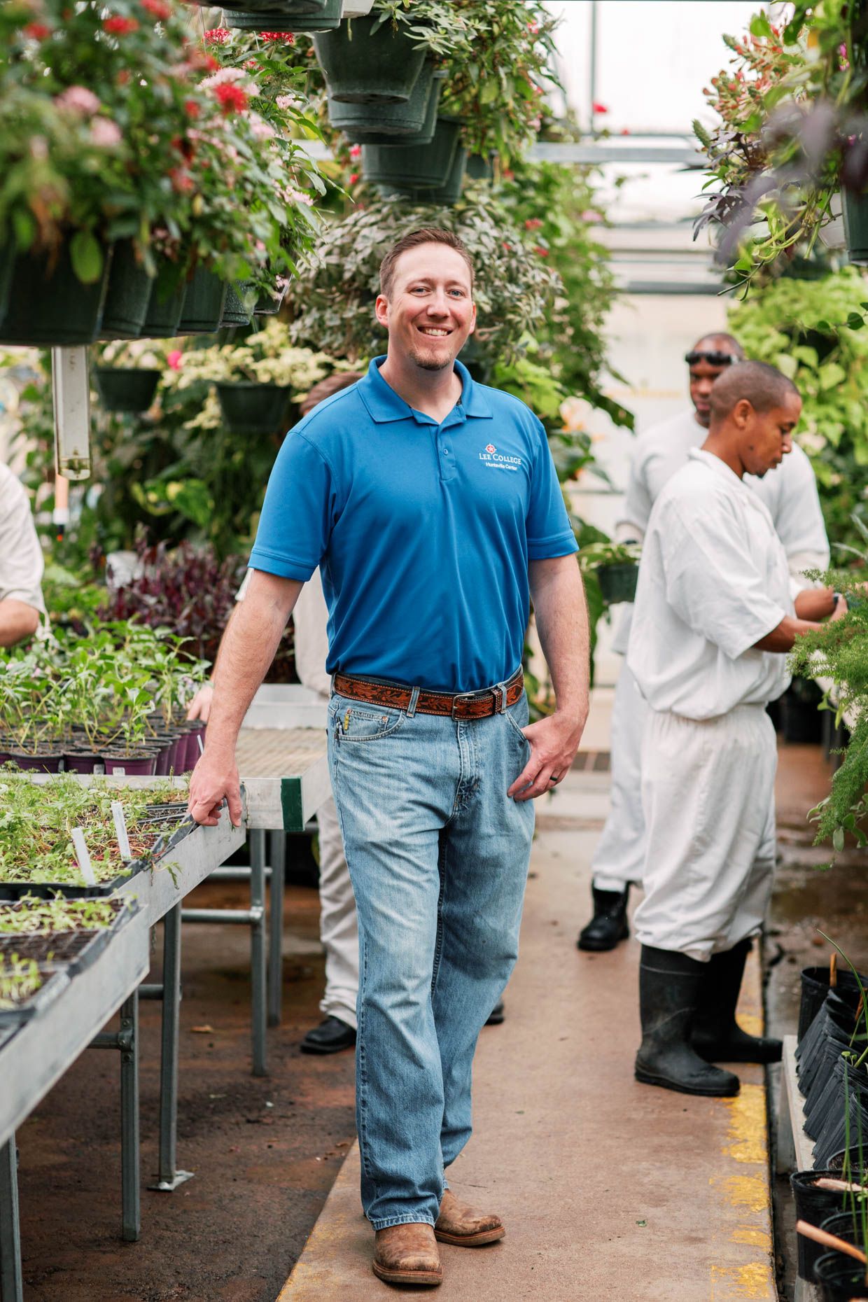 Man in blue polo shirt walks through a lush greenhouse nursery surrounded by plants and workers in white coats.