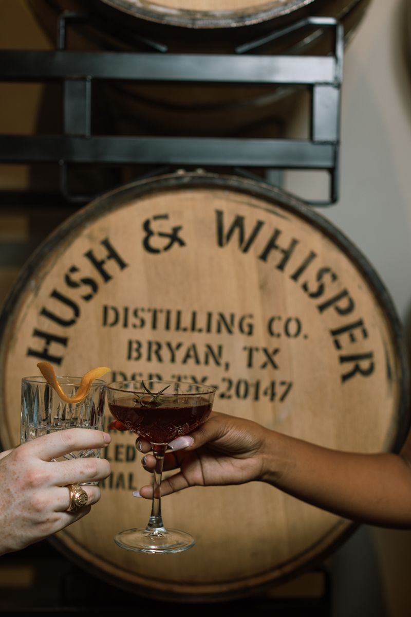 Two hands toasting cocktails in front of a Hush & Whisper Distilling Co. wooden barrel in Bryan, TX.