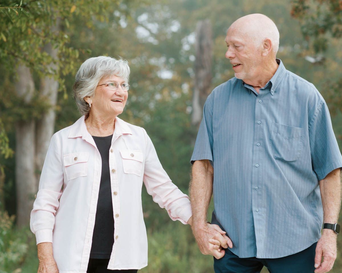 Elderly couple holding hands and smiling while taking a leisurely walk outdoors surrounded by green trees.